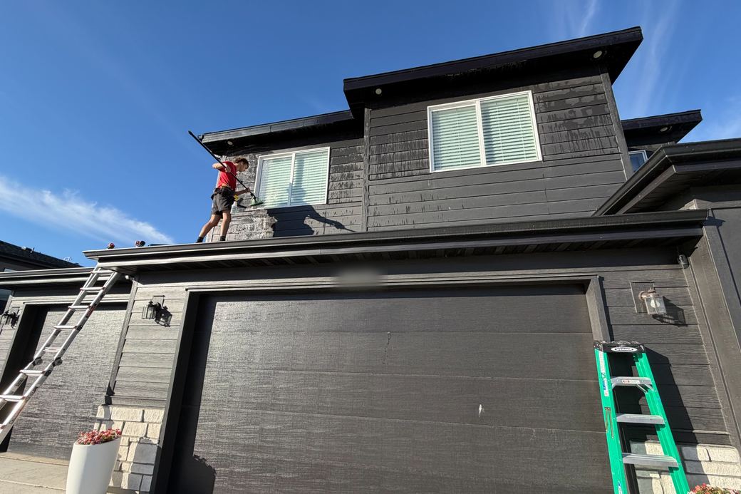 A worker using a long cleaning pole from a garage roof, with ladders set against the house.