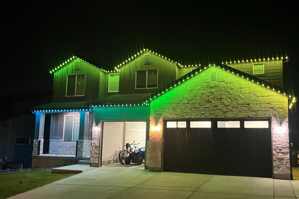 A two-story house at night with green and teal permanent lighting along the rooflines and eaves.
